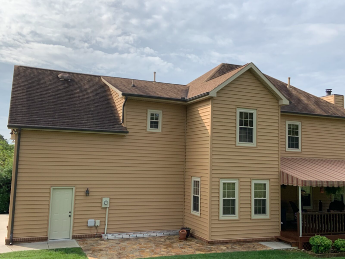 A two-story beige house with a brown roof, featuring multiple windows and a side door, stands under a partly cloudy sky. An awning over the patio adds charm to the scene. Regular roof maintenance in Knoxville ensures it remains picturesque and enduring.
