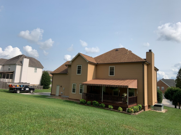 A two-story brown house with a covered porch is visible. Nearby, a pickup truck loaded with equipment for professional roof cleaning in Knoxville is parked. The well-maintained lawn and neighboring house stand out under the partly cloudy sky.