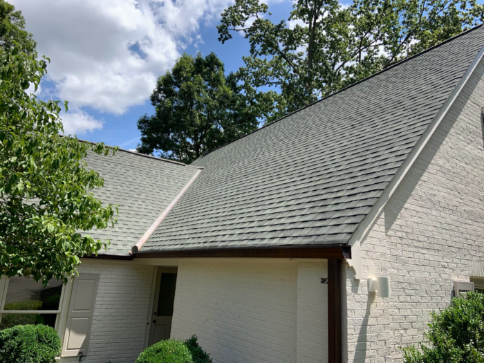 A house with a steep, gray shingled roof and white brick walls is surrounded by lush greenery and trees under a partly cloudy sky. With regular roof maintenance in Knoxville, this charming home maintains its pristine appearance.