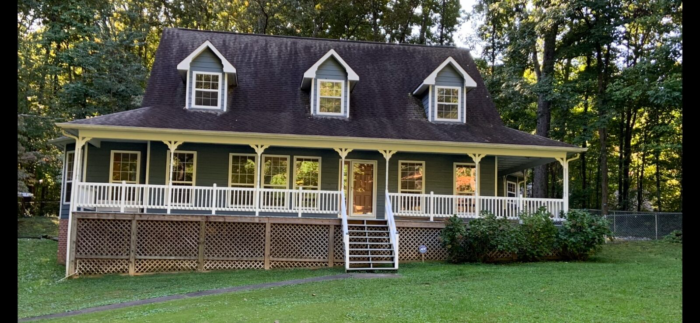 A charming two-story house with a large front porch and three dormer windows boasts a shingled roof. Nestled among trees with a grassy front yard, it benefits from regular roof maintenance, ensuring it remains pristine in the heart of Knoxville.